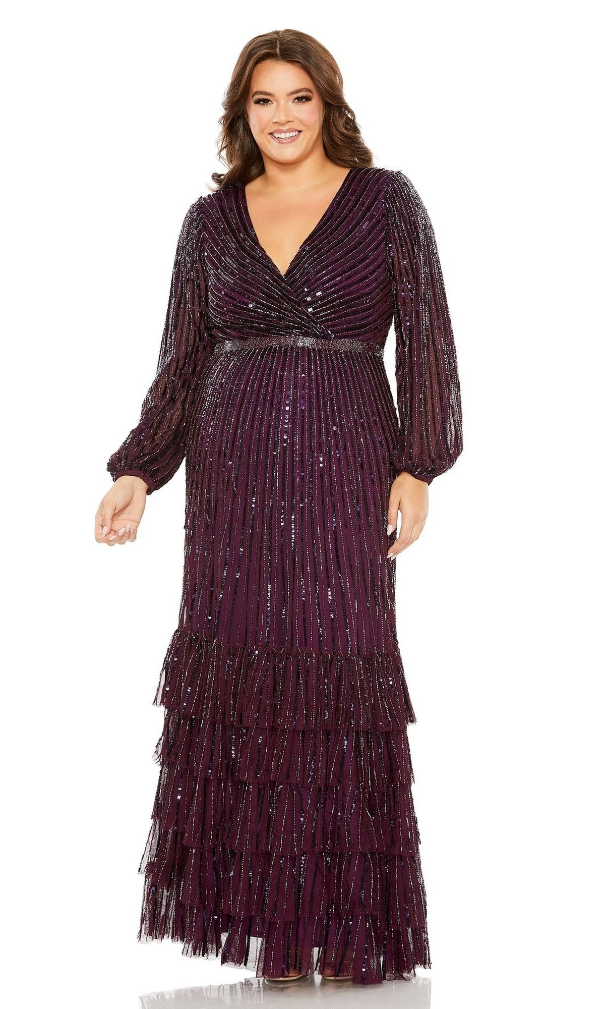 A woman smiles at the camera wearing the Long Plus-Size Formal Dress 5784 by Mac Duggal, a floor-length dark purple gown with long sleeves, a v-neckline, sequins, and tiered ruffle details.
